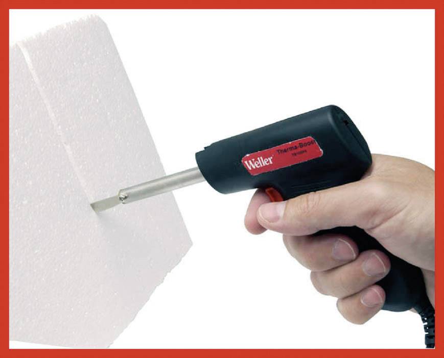 Eine Hand hält einen elektrischen Schneidstab, der durch ein Stück Styropor schneidet. Steckdosen sind sichtbar, roter Rand um das Bild.