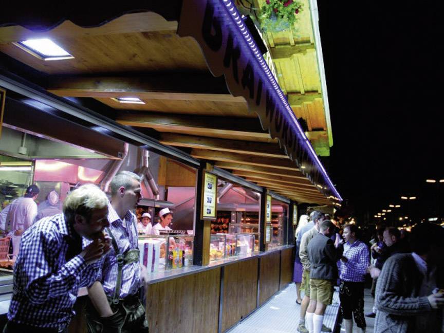 Menschen stehen in Tracht bei einem belebten Nachtmarkt vor einem beleuchteten Imbissstand. Essen wird zubereitet und verkauft.