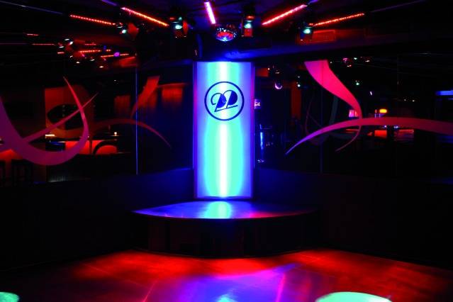 Eine bunte Tanzfläche in einem Club mit Neonlichtern und einem beleuchteten Podium in der Mitte.