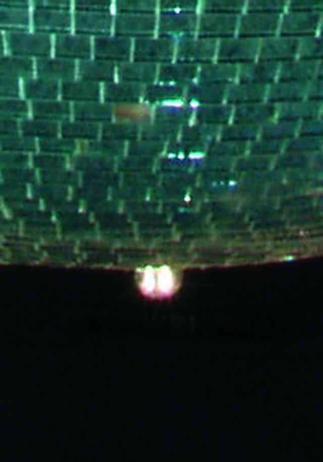 Eine leuchtende Kugel hängt von einer mit Spiegelplättchen bedeckten Decke, die Licht reflektieren. Dunkler Hintergrund.