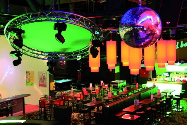 Eine bunte Bar mit neonbeleuchtetem Ambiente, Discolicht und Spiegelkugel. Reihen von Sitzplätzen und eine gut beleuchtete Theke.