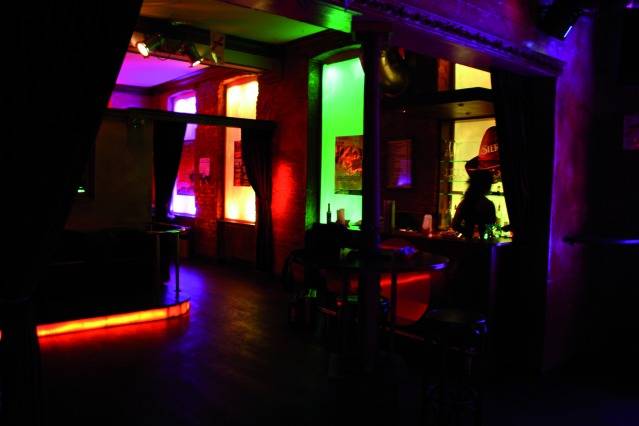 Ein schwach beleuchteter Raum in einer Bar oder einem Club mit bunten Lichtern an den Wänden, einer Bar und Barhocker im Vordergrund.