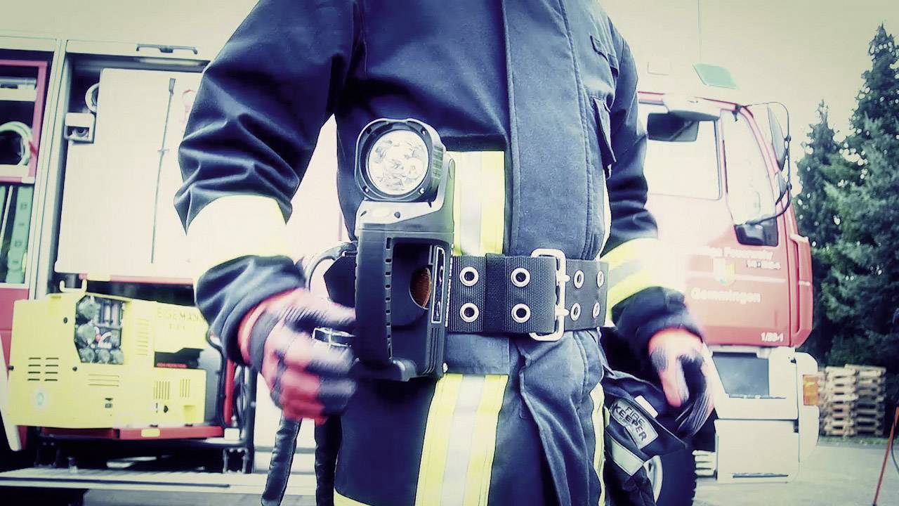 Ein Feuerwehrmann in voller Einsatzkleidung steht vor einem Feuerwehrauto. Er trägt eine Taschenlampe an einem Gürtel befestigt.