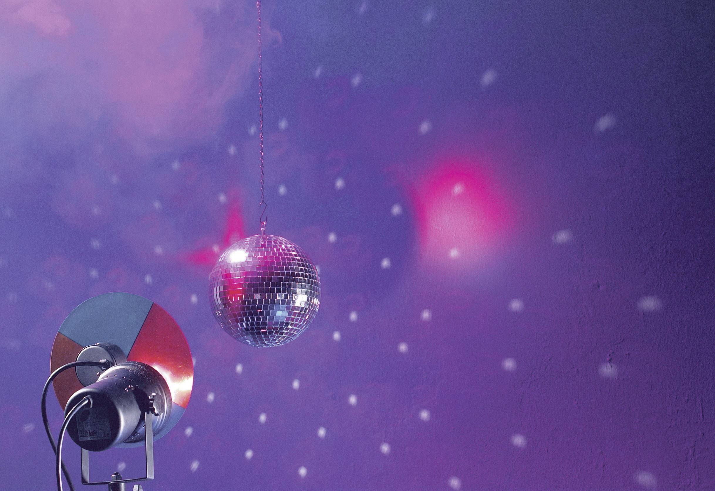 Discokugel hängt von der Decke. Eine bunte Lampe leuchtet darauf und erzeugt bunte Lichtpunkte auf einer lila Wand.