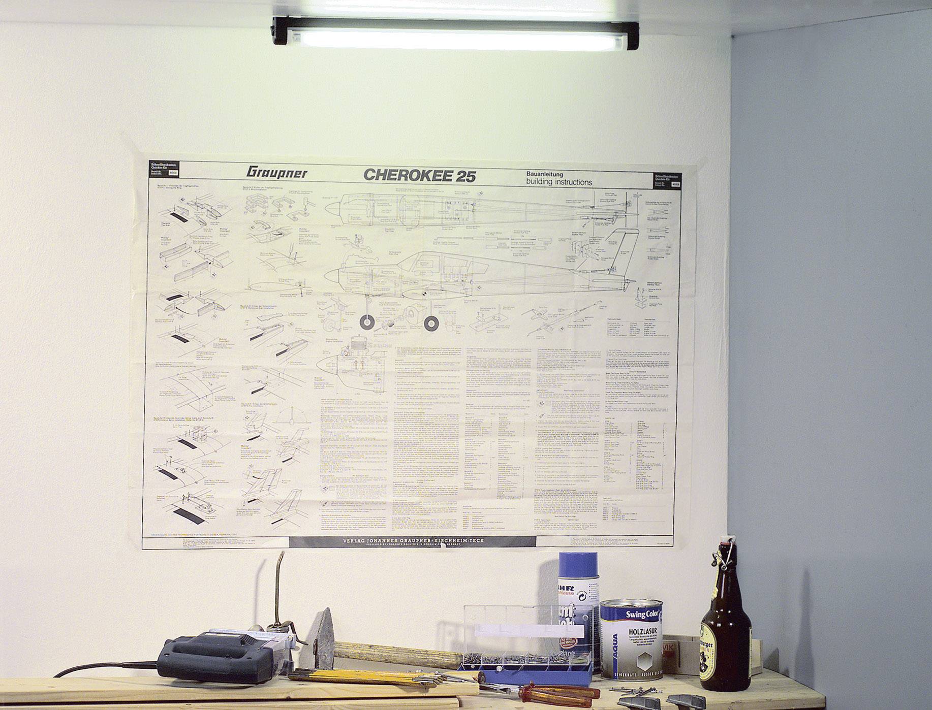 Bauplan eines Modellflugzeugs hängt an der Wand; davor Werkzeuge und Materialien auf einem Tisch, darunter Klebstoff und leere Flasche.