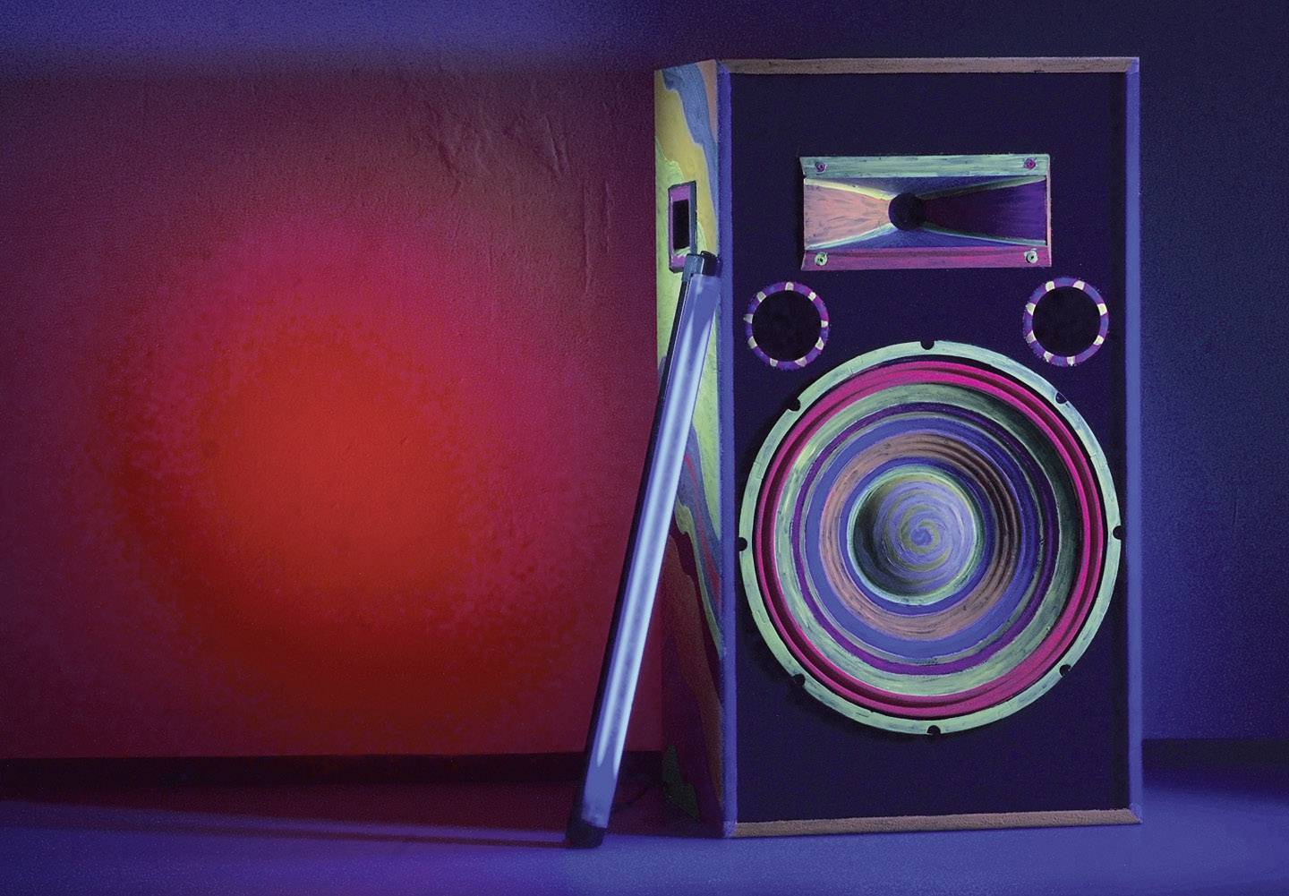 Ein bunter Lautsprecher im Stil der 80er Jahre steht vor einer rot-blauen Wand. Eine lange Röhre lehnt an der Seite des Lautsprechers.