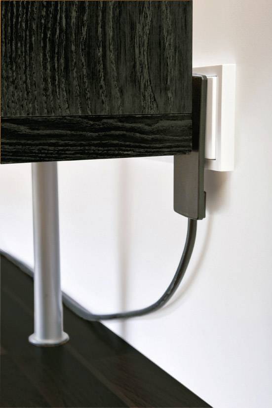 'Ein schwarzes Möbelstück steht dicht an einer weißen Wand. Ein schwarzes Kabel ist in eine Steckdose an der Wand eingesteckt.'