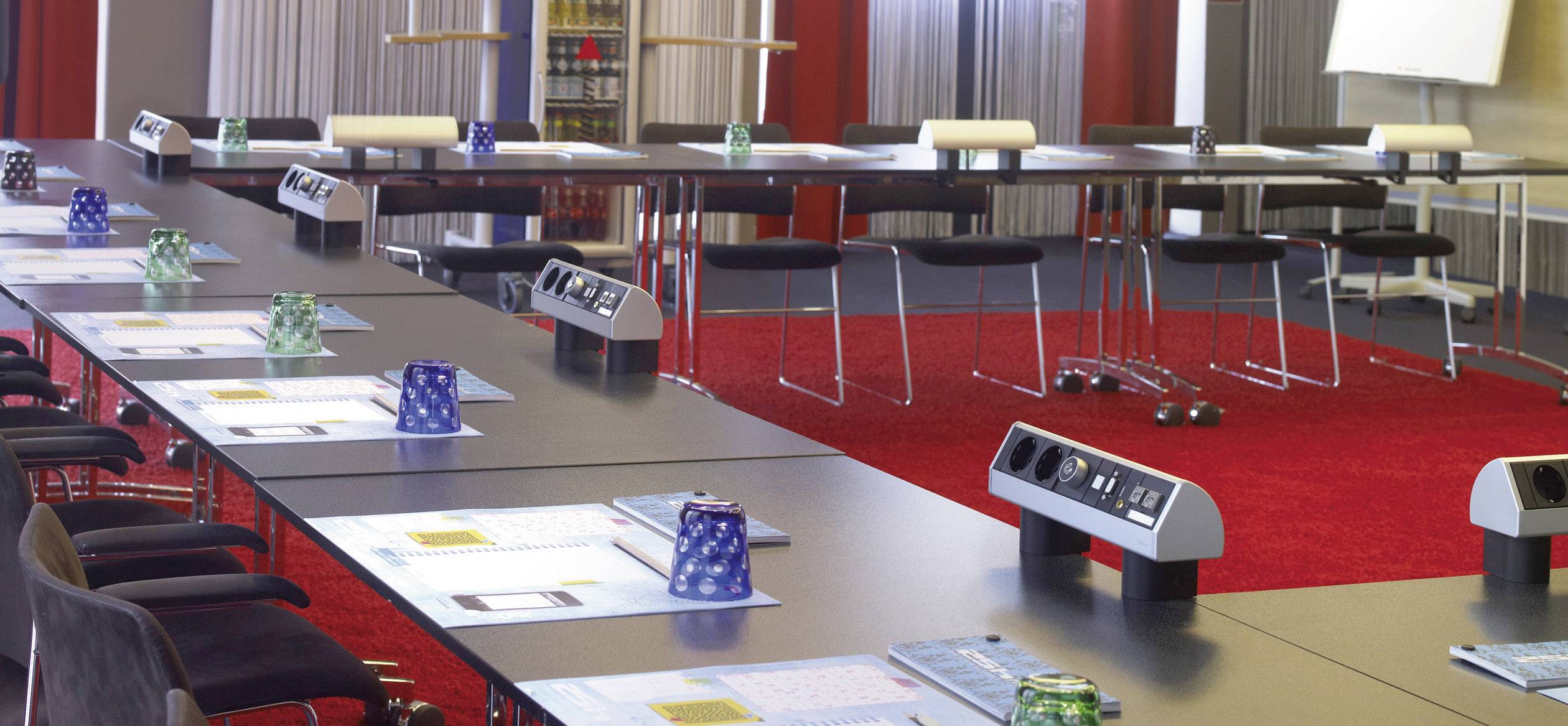 Konferenzraum mit mehreren rechteckigen Tischen, Stühlen und Schreibmaterialien. Die Tische sind U-förmig angeordnet auf rotem Teppich.