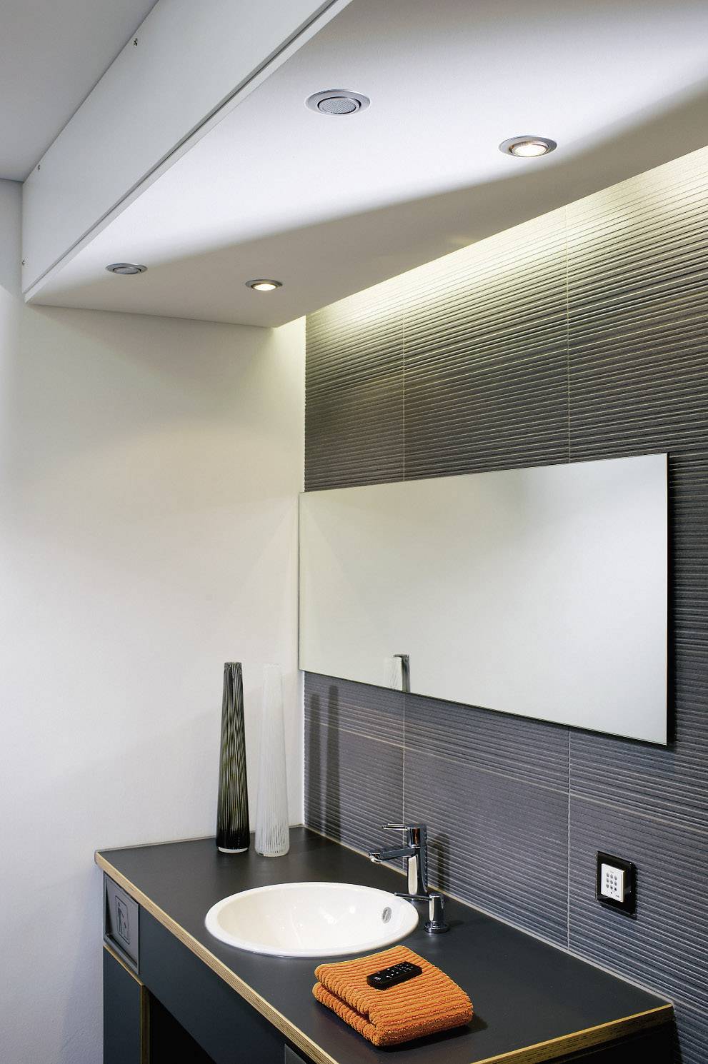 Modernes Badezimmer mit Waschbecken, Spiegel, Vase und orangefarbenem Handtuch. Dezente Beleuchtung mit eingebauten Leuchten an der Decke.