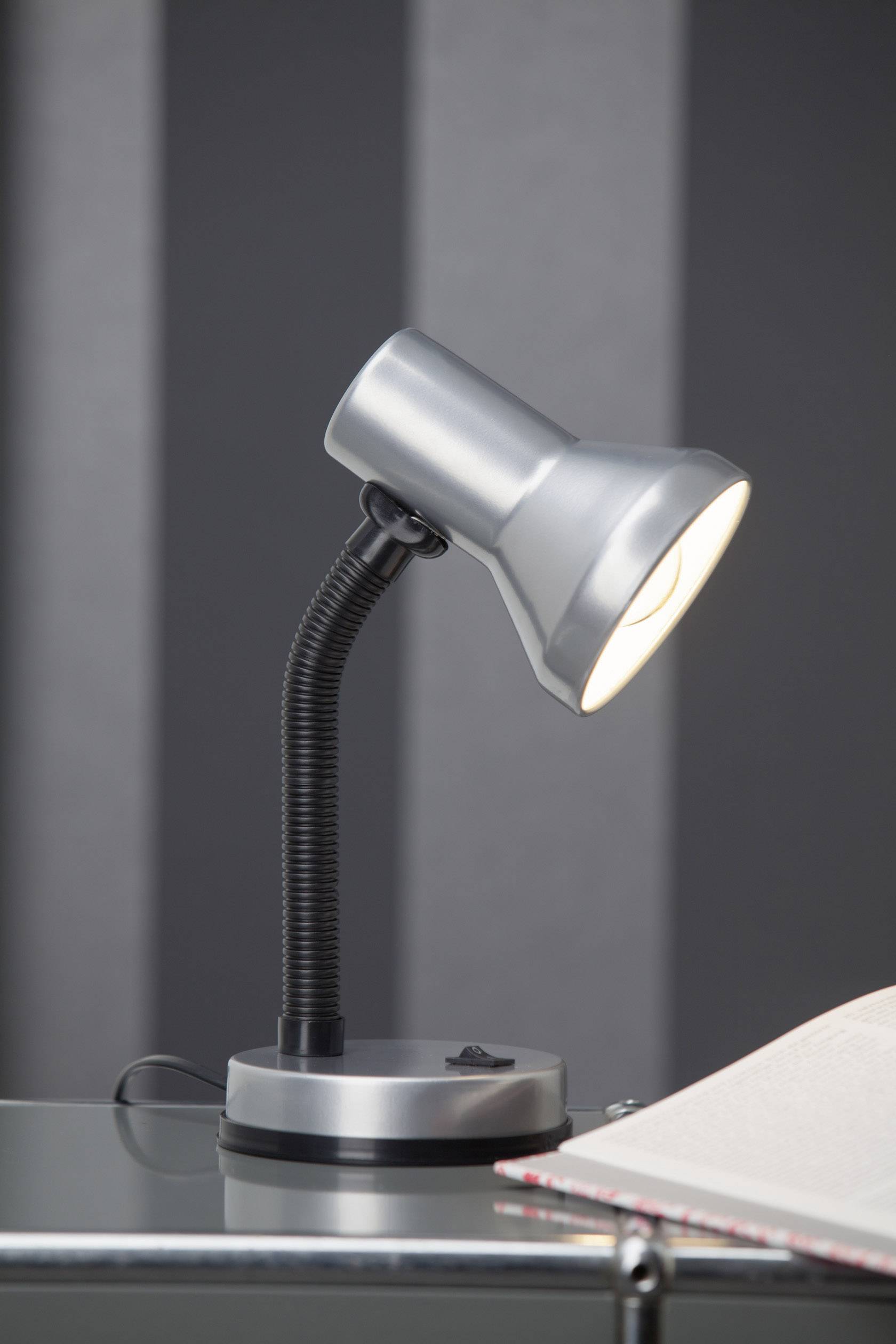 Eine silberne Tischlampe leuchtet auf ein offenes Buch auf einem Glastisch, vor einem gestreiften grauen Hintergrund.