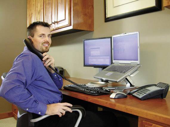 Mann in blauem Hemd telefoniert in einem modernen Büro, Blick auf zwei Computerbildschirme.
