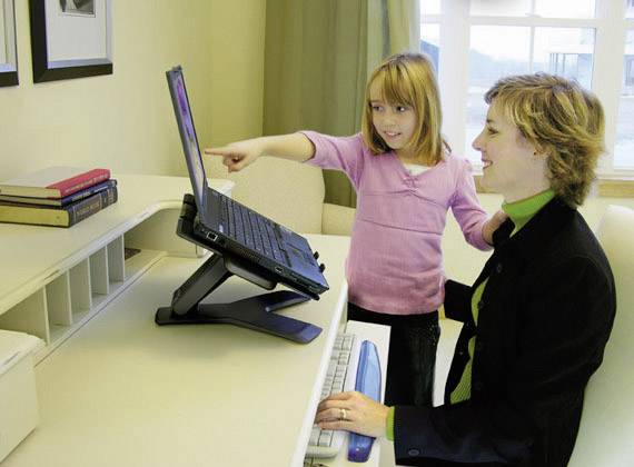 Ein Kind zeigt einer Frau auf einem Laptop-Bildschirm etwas. Beide lächeln, die Frau sitzt an einem Schreibtisch mit Büchern.