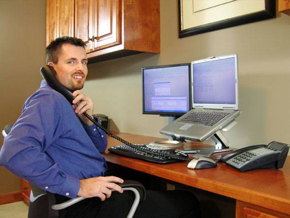 Ein Mann sitzt an einem Schreibtisch und telefoniert. Vor ihm stehen zwei Computerbildschirme. Er trägt ein blaues Hemd und lächelt.