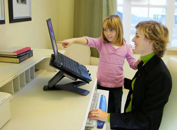 Eine Frau sitzt am Schreibtisch, während ein Mädchen auf den Laptopbildschirm zeigt. Bücher liegen daneben. Helle Raumbeleuchtung.