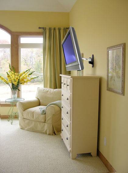 Ein Wohnzimmer mit einem beigen Sessel, einer Kommode, darauf montiert ein Flachbildfernseher. Links ein Tisch mit gelben Blumen.