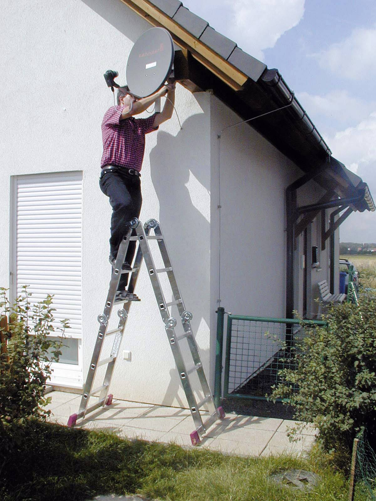 Eine Person steht auf einer Leiter und montiert eine Satellitenschüssel an der Dachkante eines Hauses.