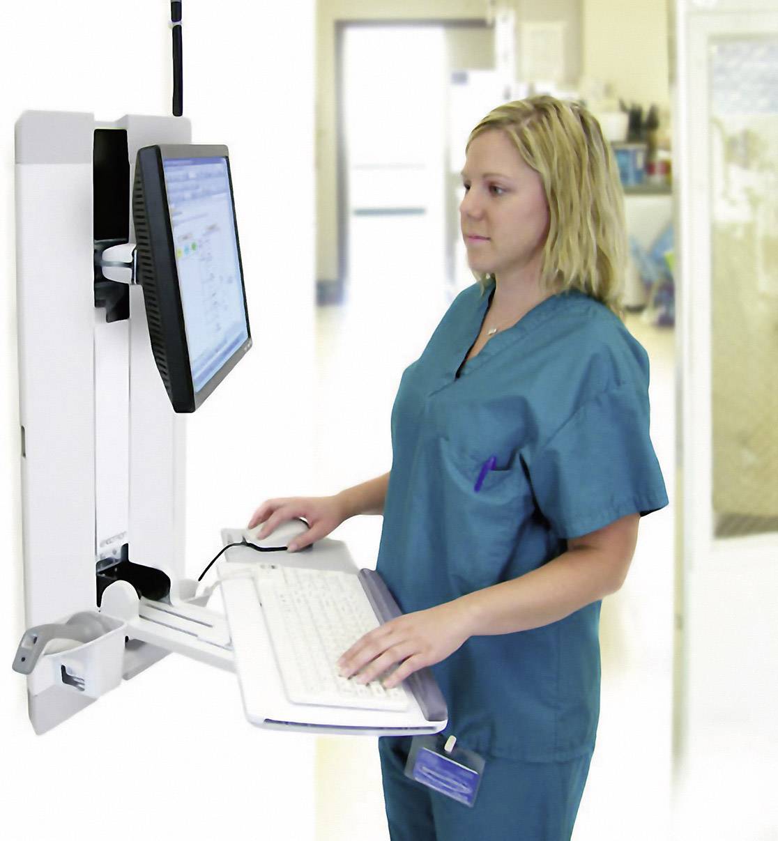 Eine Frau in Krankenhauskleidung steht an einem Computerarbeitsplatz, tippt auf der Tastatur und schaut auf den Bildschirm.