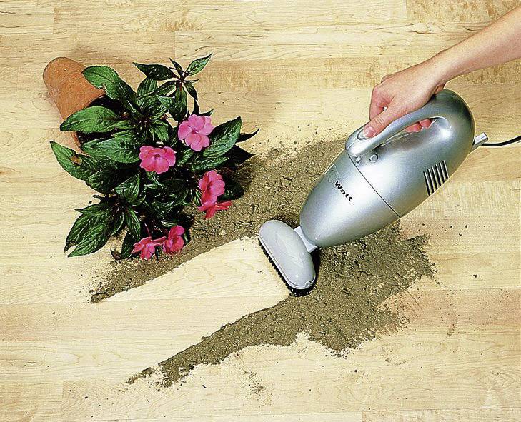 Handstaubsauger entfernt verschüttete Erde vom Boden neben umgekipptem Blumentopf mit rosa Blüten.