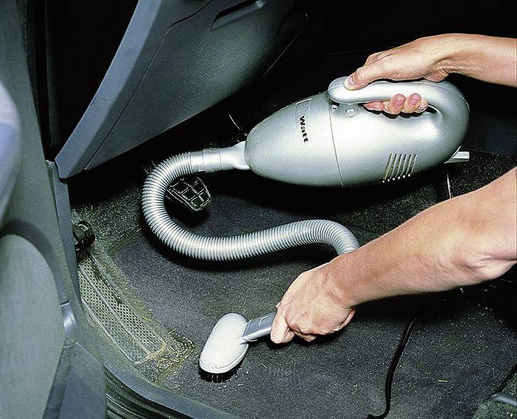 Person saugt Fußmatte eines Autos mit einem tragbaren Staubsauger ab. Die Szene zeigt gründliche Reinigung.