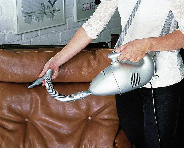 Eine Person hält einen tragbaren Staubsauger und reinigt ein Ledersofa.