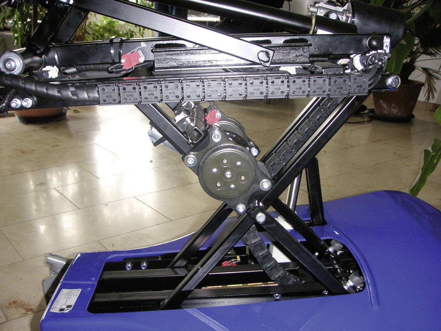 Ein blauer, verstellbarer Hebemechanismus mit schwarzem Rahmengestell und Elektromotor ist auf einem hellen Boden montiert.