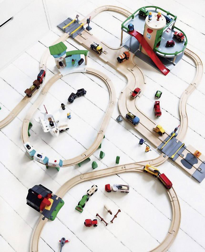 Holzeisenbahn-Set auf weißem Boden, mit Schienen, Fahrzeugen, Figuren und Gebäuden. Zentrale grüne Plattform und mehrere Verbindungswege.