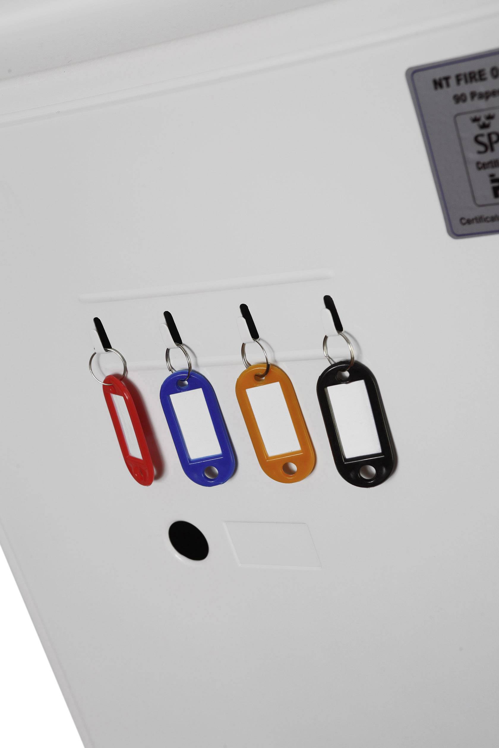 Vier bunte Schlüsselanhänger (rot, blau, orange, schwarz) hängen an Haken an einer weißen Wand. Oben rechts ein Schild.