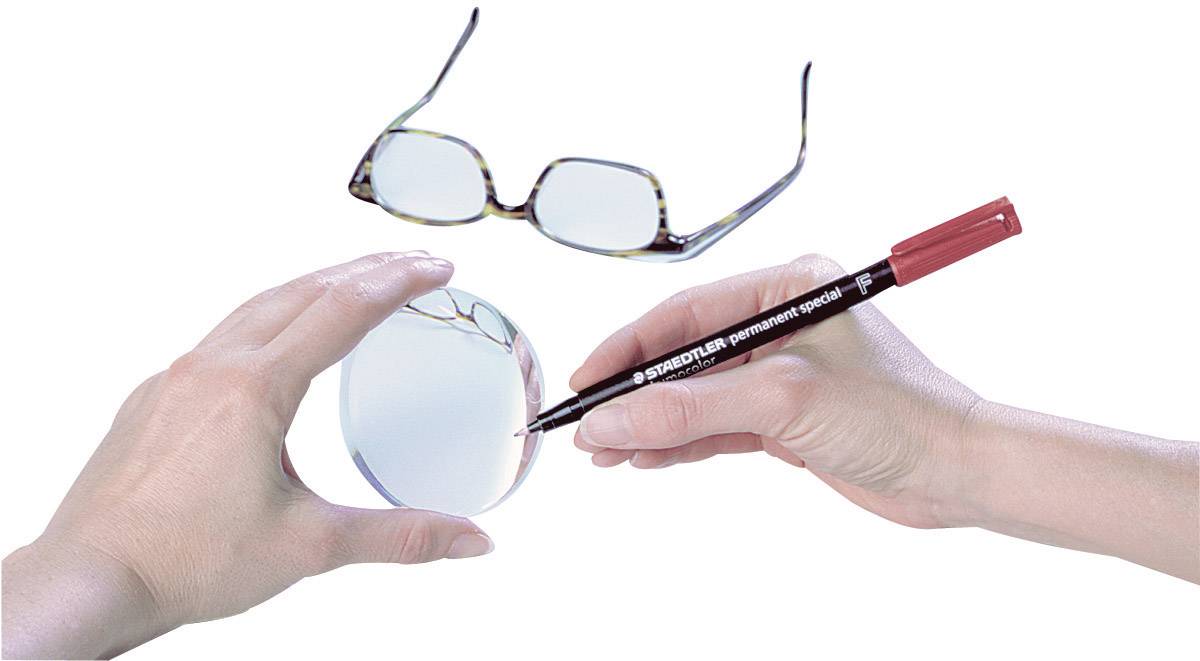 Eine Hand hält eine Brillelinse, während eine andere Hand mit einem roten Stift eine Markierung darauf macht. Im Hintergrund liegt eine Brille.