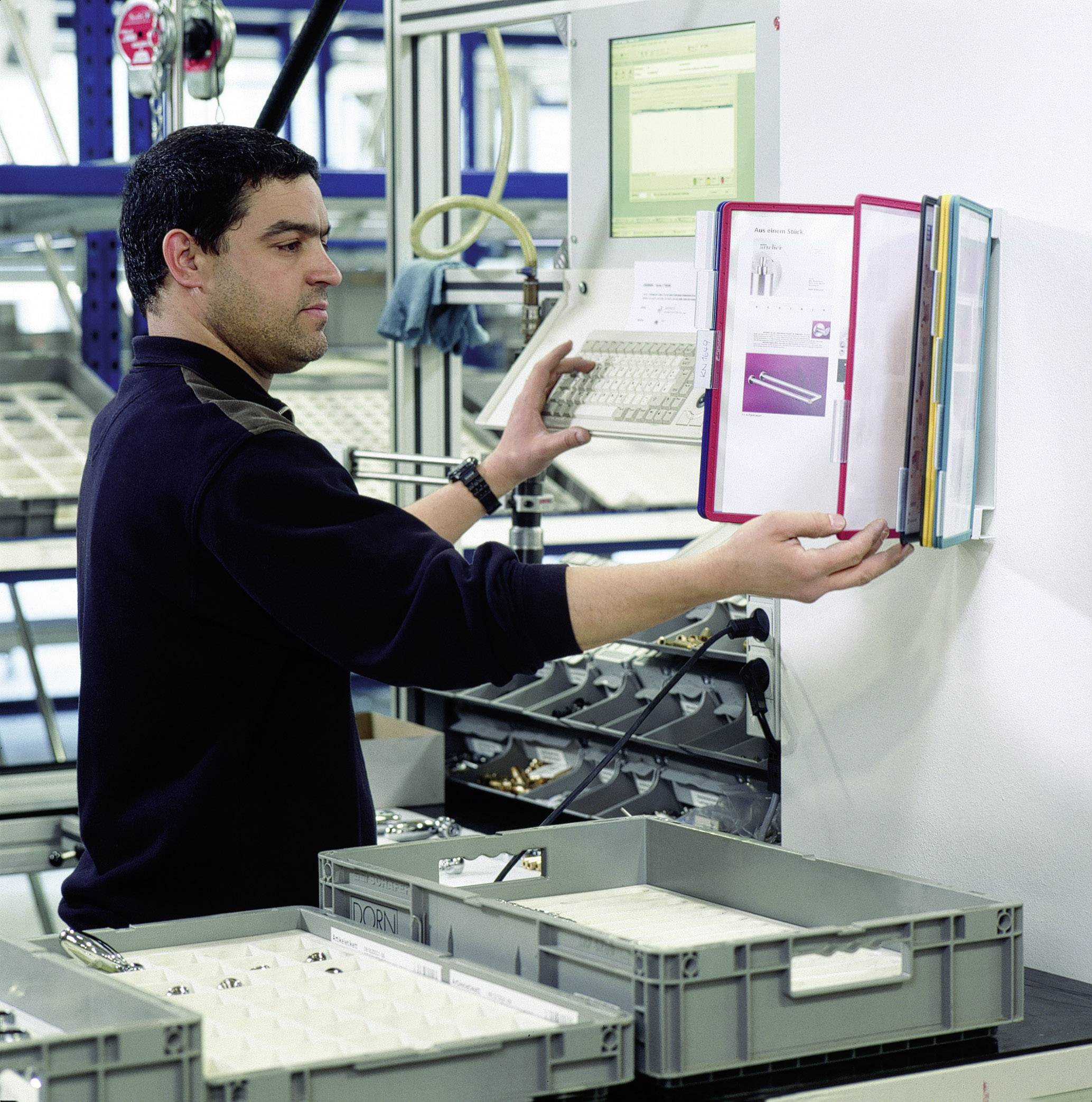 Ein Mann arbeitet in einer Fabrikumgebung, bedient einen Computer und überprüft Dokumente in einem Aktenordner, vor mehreren Ablagekisten.