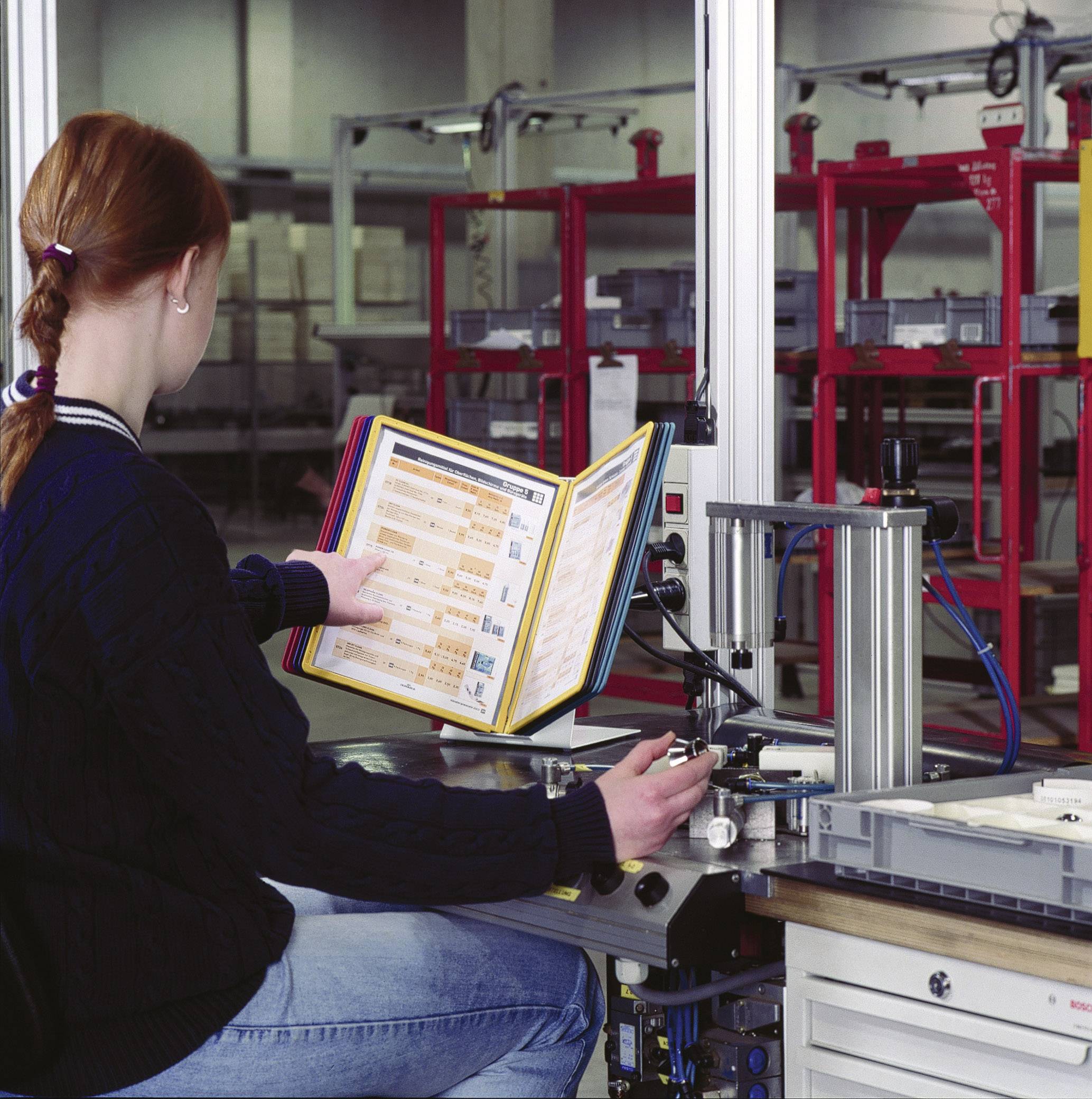 Eine Person sitzt an einem Arbeitstisch in einer Produktionshalle und überprüft ein geöffnetes Handbuch. Im Hintergrund sind Maschinen zu sehen.