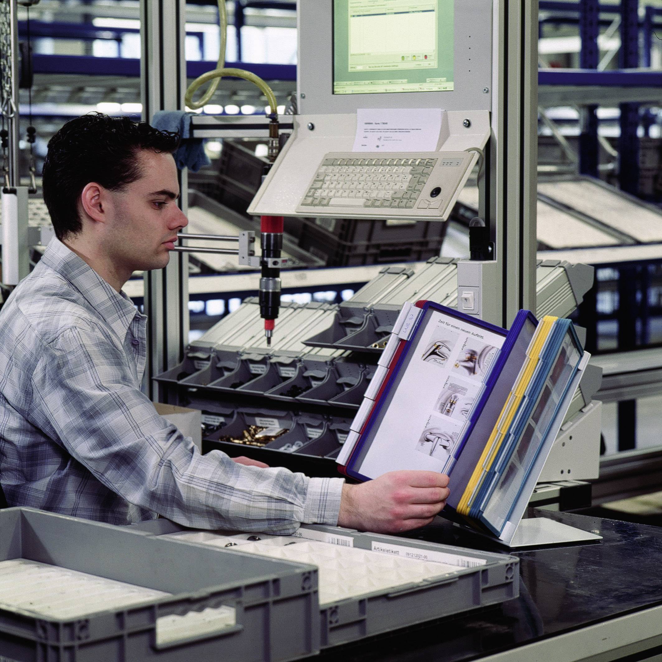 Ein Mann steht in einem modernen Lager. Er blättert durch einen Ordner und arbeitet an einem Computer, der an einem Regal angebracht ist.