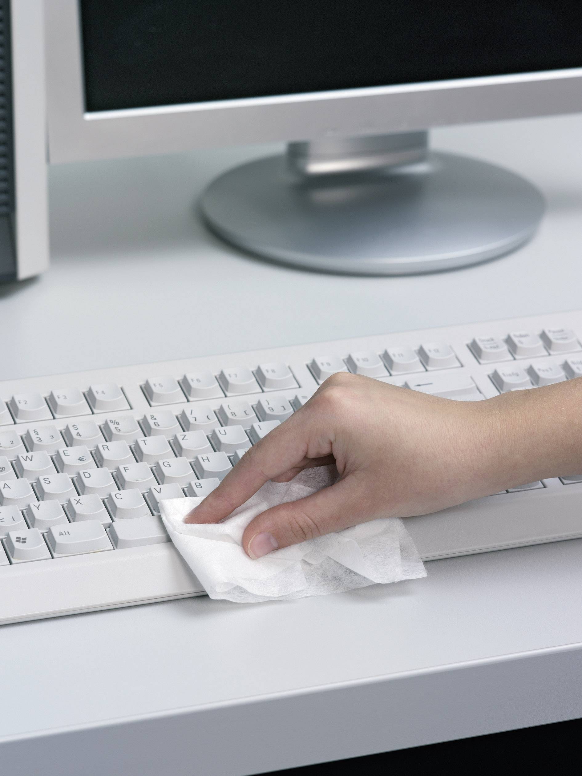 Hand reinigt eine Computertastatur mit einem weißen Tuch vor einem Monitor.