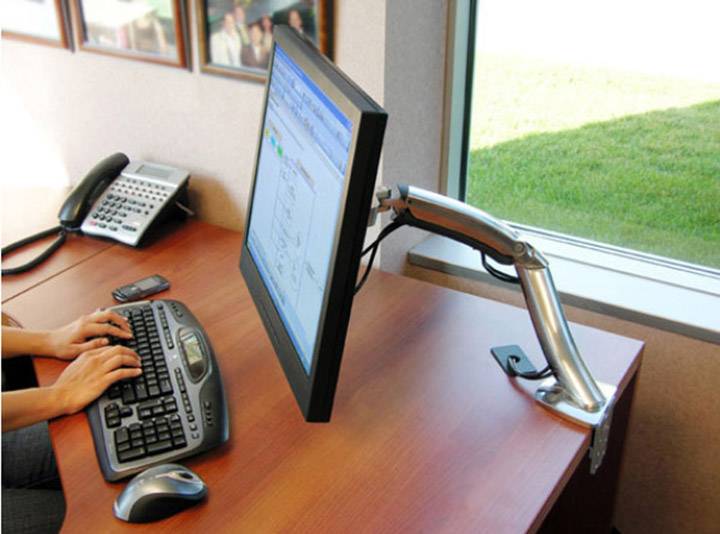 Eine Person arbeitet an einem Computer mit einem höhenverstellbaren Monitorarm. Der Arbeitsplatz ist neben einem Fenster mit Blick nach draußen platziert.