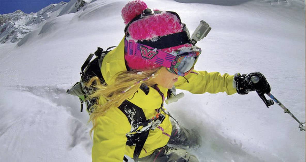 Eine Person in gelber Jacke fährt auf Skiern durch tiefen Schnee einen Berg hinunter. Sie trägt eine pinke Mütze und Skibrille.