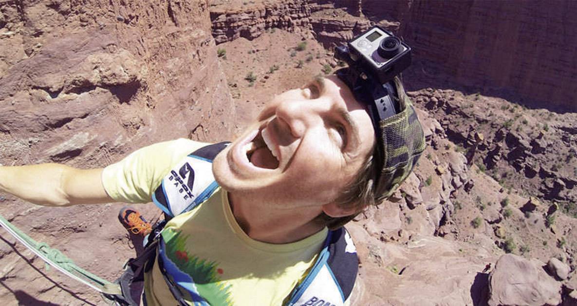 Eine Person klettert an einer steilen Felswand und trägt eine Kamera auf dem Kopf. Sie wirkt fröhlich und konzentriert.