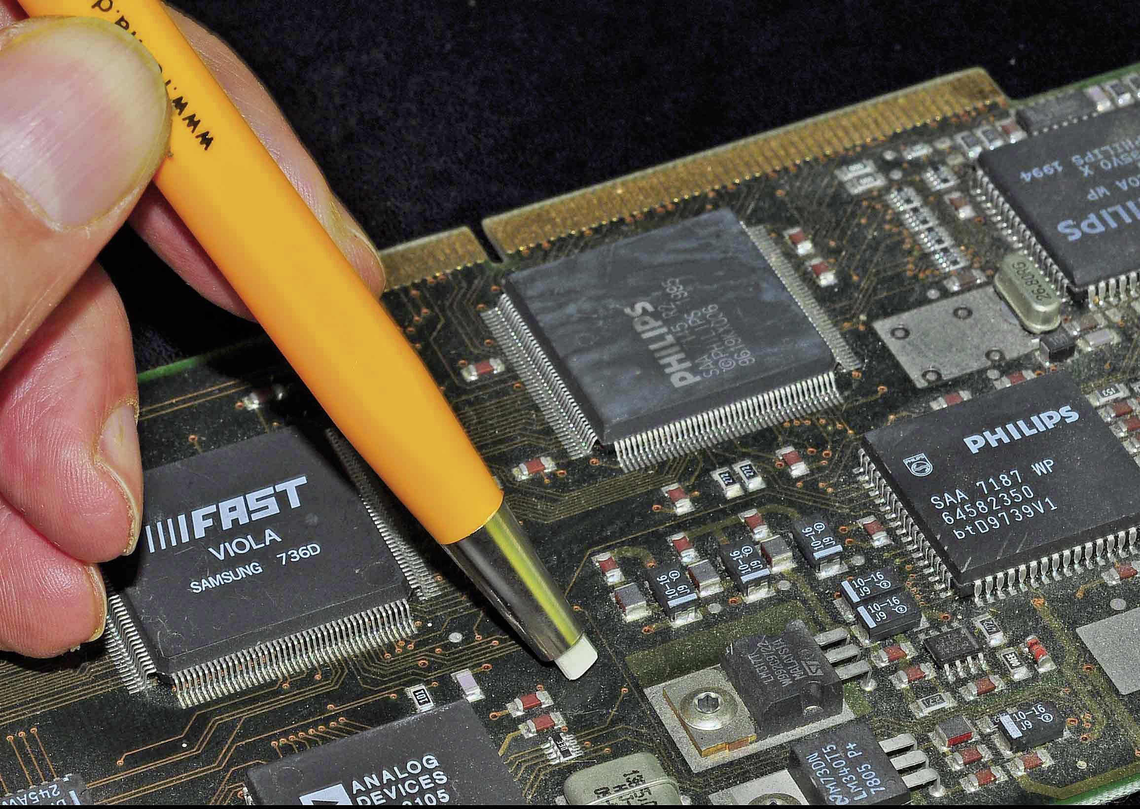 Hand zeigt mit Stift auf Computerplatine mit mehreren integrierten Schaltkreisen, darunter 'PHILIPS' und 'ANALOG DEVICES'.