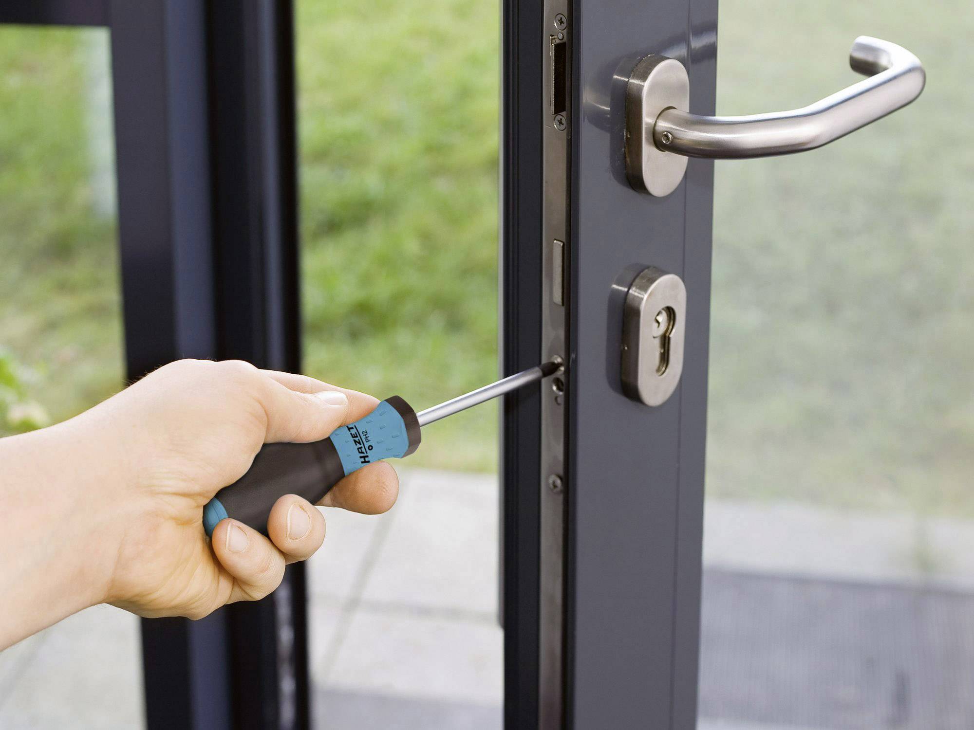 Eine Hand benutzt einen Schraubendreher, um ein Schloss in einem Türrahmen zu reparieren. Draußen ist unscharfes Grün zu sehen.