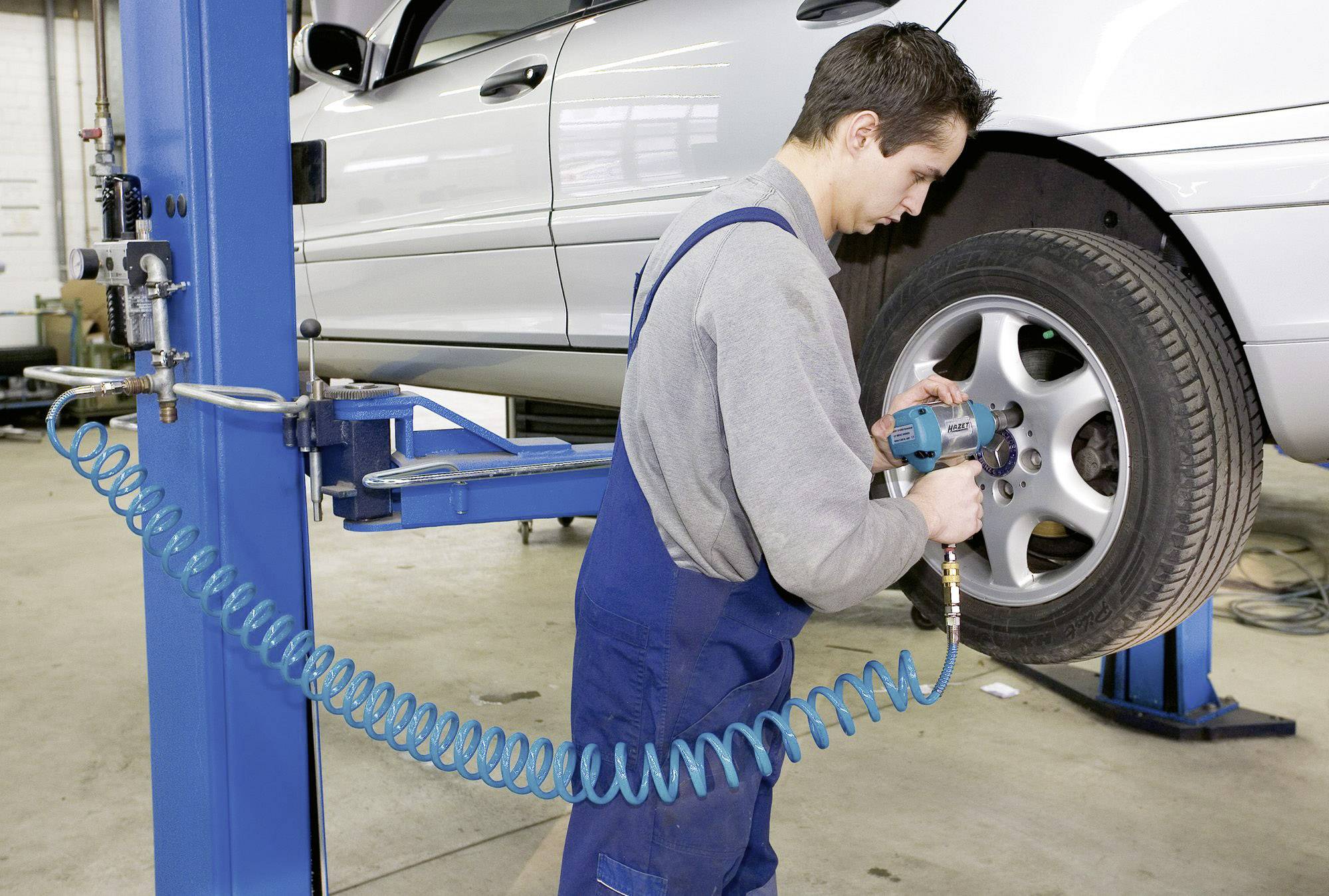 Ein Mechaniker in blauer Latzhose wechselt einen Reifen an einem angehobenen Auto in einer Werkstatt.
