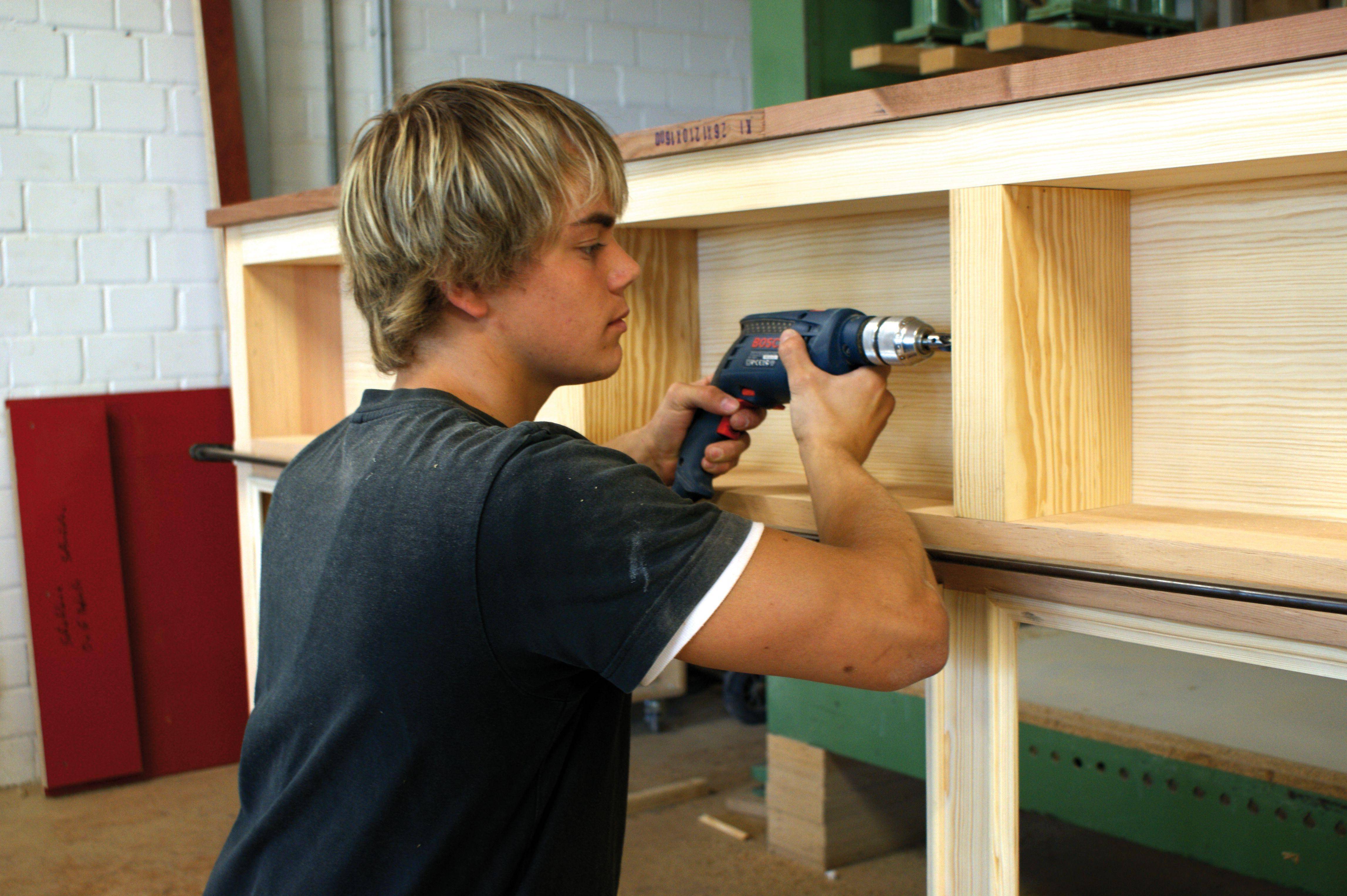 Ein junger Mann benutzt einen Bohrer, um ein Holzregal zu montieren. Er konzentriert sich auf seine Arbeit in einer Werkstattumgebung.