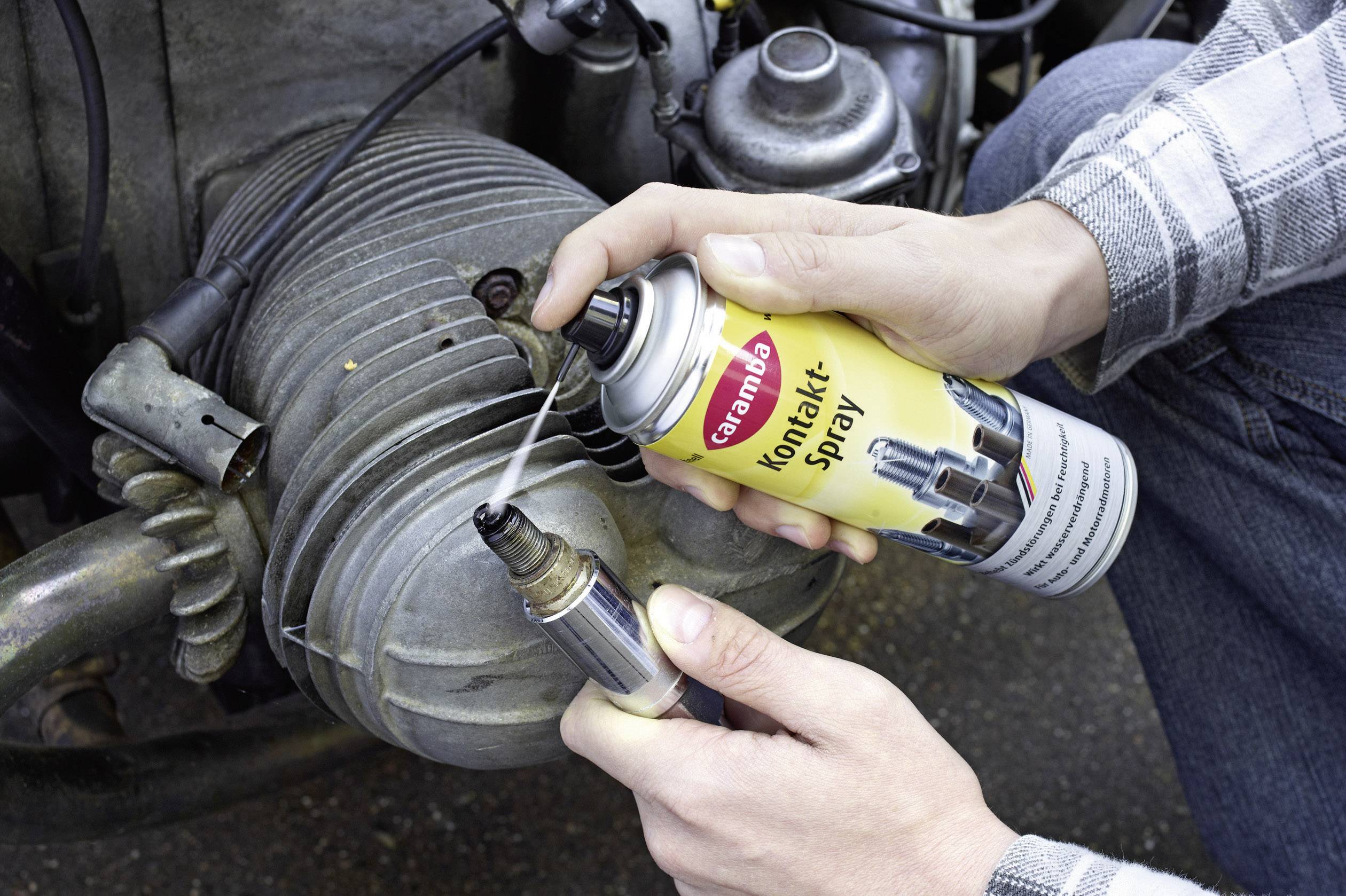 Eine Person sprüht Kontaktspray auf eine Zündkerze vor einem Motorblock, um die elektrische Leitfähigkeit zu verbessern.