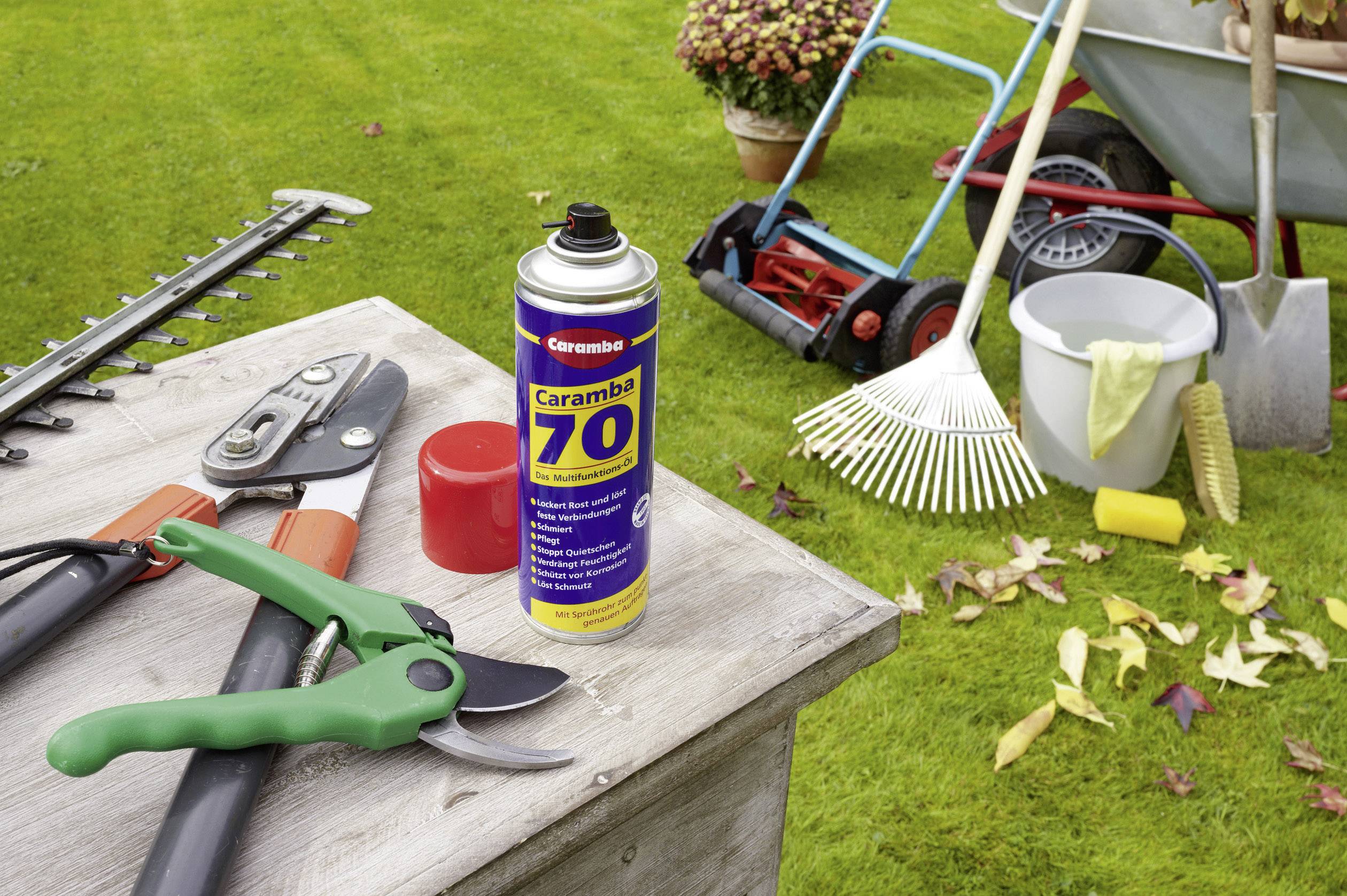Gartenarbeitsplatz mit Astschere, Spraydose, Rechen und anderen Werkzeugen auf einem Holztisch. Hintergrund zeigt Rasenmäher und Schubkarre auf grünem Rasen.