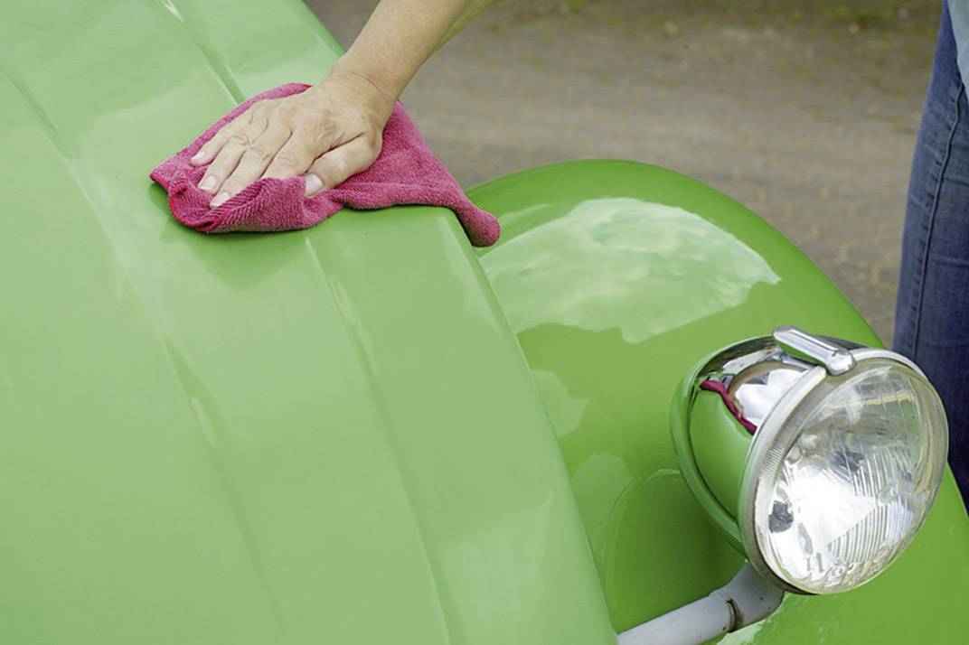 Eine Hand poliert mit einem roten Tuch die Motorhaube eines grünen Autos, betont durch ein glänzendes Scheinwerferdetail.