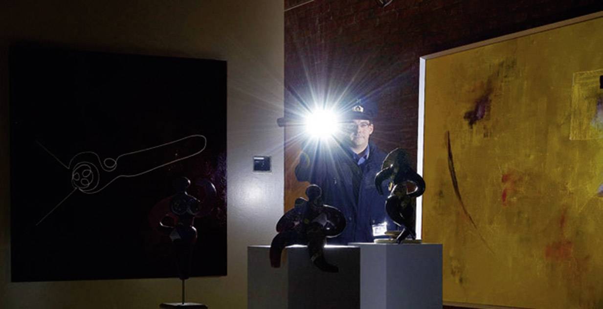 Person in einem dunklen Raum leuchtet mit einer Taschenlampe auf Kunstwerke. Kunstwerke bestehen aus abstrakten Gemälden und Skulpturen.