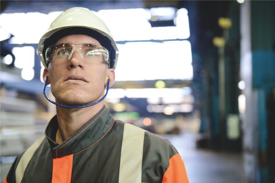 Mann mit Helm, Schutzbrille und 3M Bügelgehörschutz bei der Arbeit in einer Industriehalle.