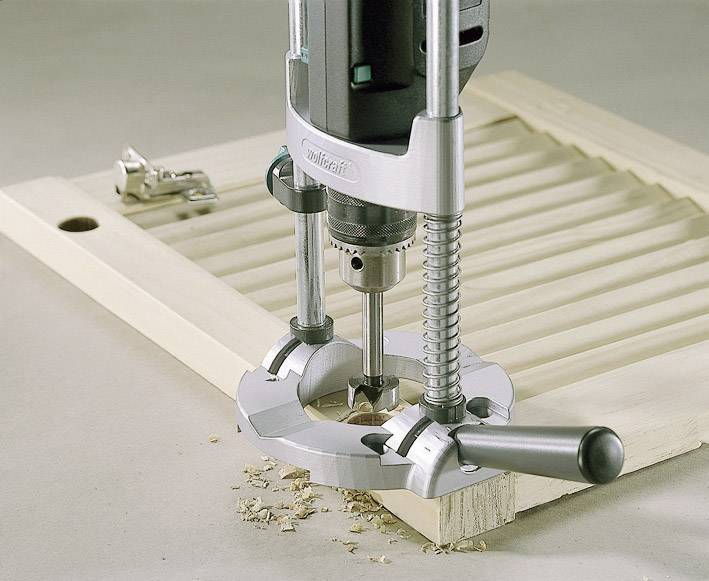 Eine Fräse bearbeitet ein Holzstück, um eine Vertiefung zu erstellen. Späne liegen auf der Oberfläche. Im Hintergrund ein Scharnier.