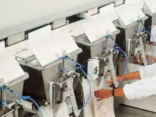 Produktionslinie mit mehreren maschinellen Stationen, an denen Arbeiter in weißer Schutzkleidung große Papierbeutel füllen.