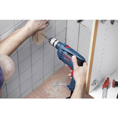 Eine Person benutzt eine blaue Bohrmaschine, um ein Holzstück an eine geflieste Wand zu schrauben. Schrauben und Werkzeug sind daneben zu sehen.