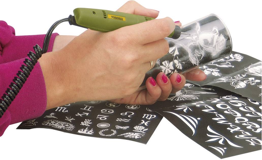 Person graviert mit einem elektrischen Gravurstift Muster auf ein Glas. Verschiedene Gravurvorlagen und Symbole liegen daneben.