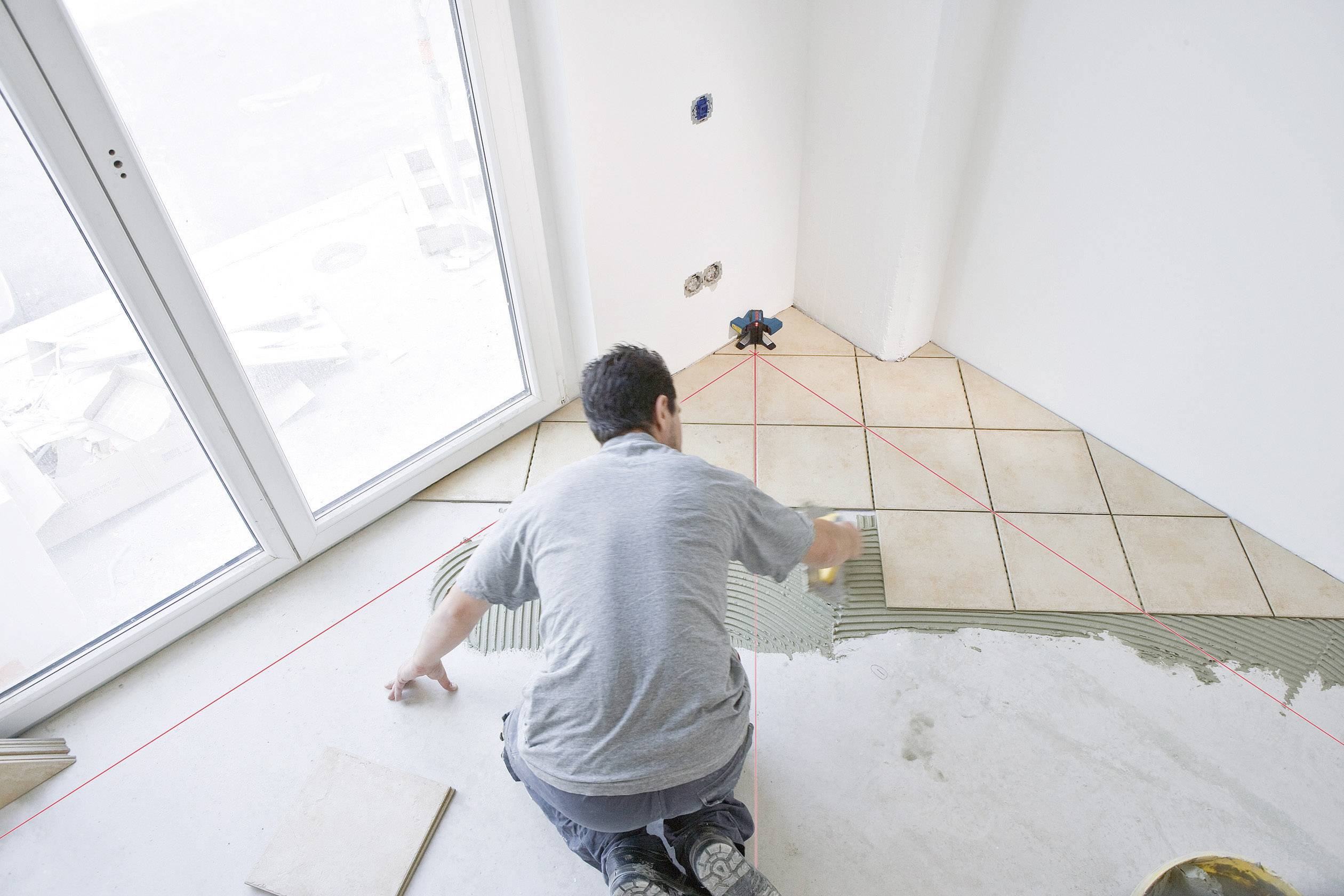 Ein Mann verlegt in einem Raum mit großen Fenstern Fliesen auf einem Boden. Rote Linien helfen bei der Ausrichtung der Fliesen.