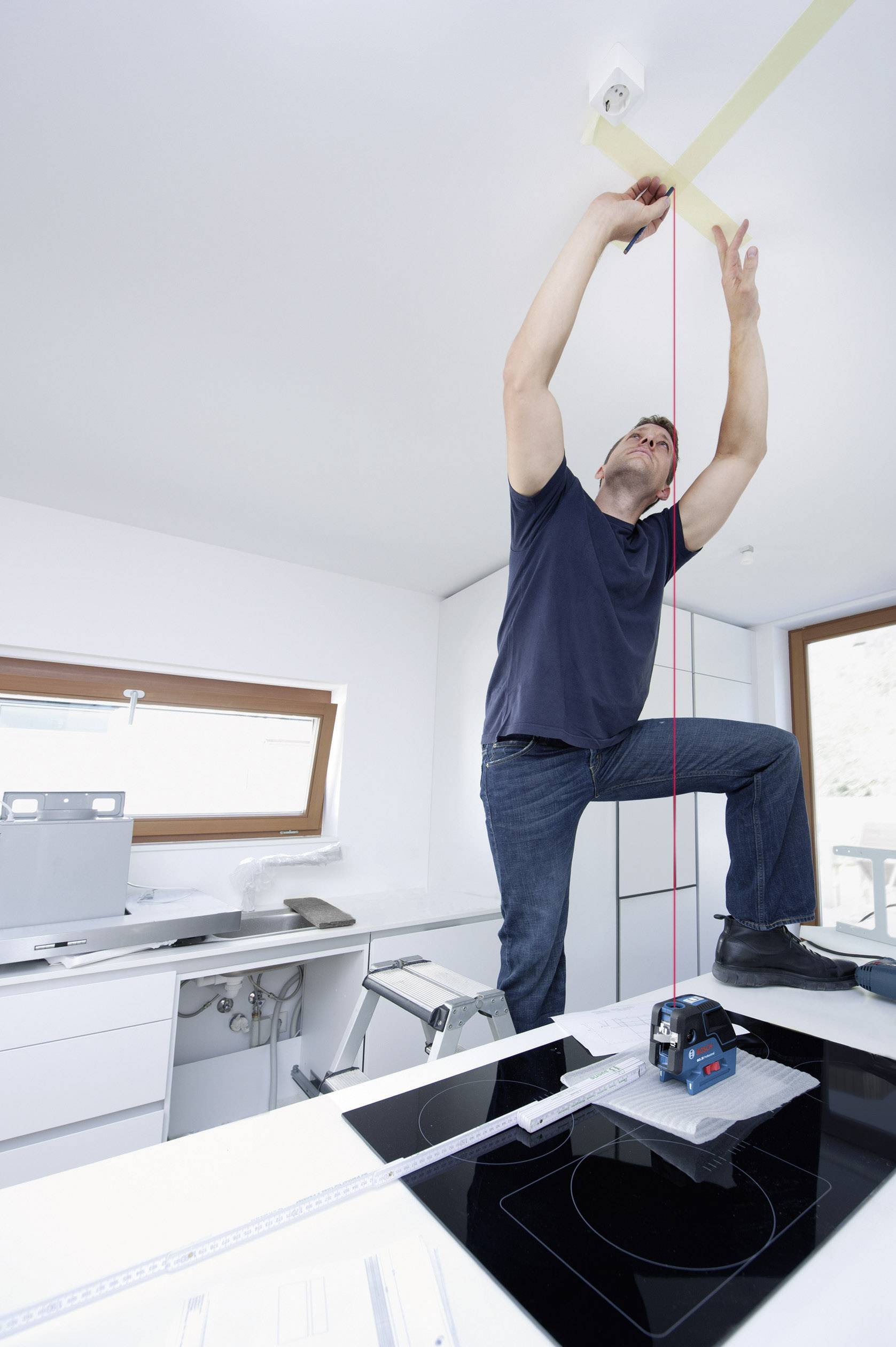 Ein Handwerker befestigt eine Lampe an der Decke in einer modernen Küche. Er steht auf einer Leiter und nutzt Werkzeug zur Installation.